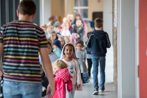 Leerkracht met rug naar lens voor een rij kleuters. Eentje kijkt lachend naar fotograaf.