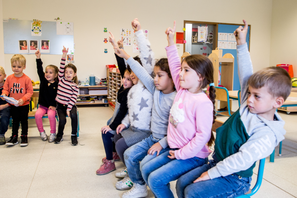 Gretige Kleuters Op Een Bank In De Klas Met Handjes In De Lucht
